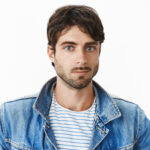 headshot of attractive young hispanic man with blue eyes and beard in denim jacket over striped t shirt looking at camera with nice friendly expression, smiling as enjoying view from new apartment